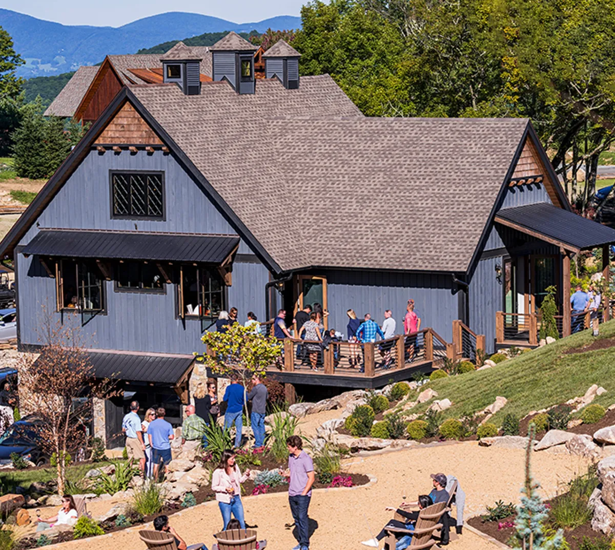 A welcoming exterior shot of the house featuring a grey facade, wooden deck, landscaped garden, and people enjoying the outdoor space. The scene exudes a sense of arrival and community, highlighting the home as a destination for travelers to relax and recharge.