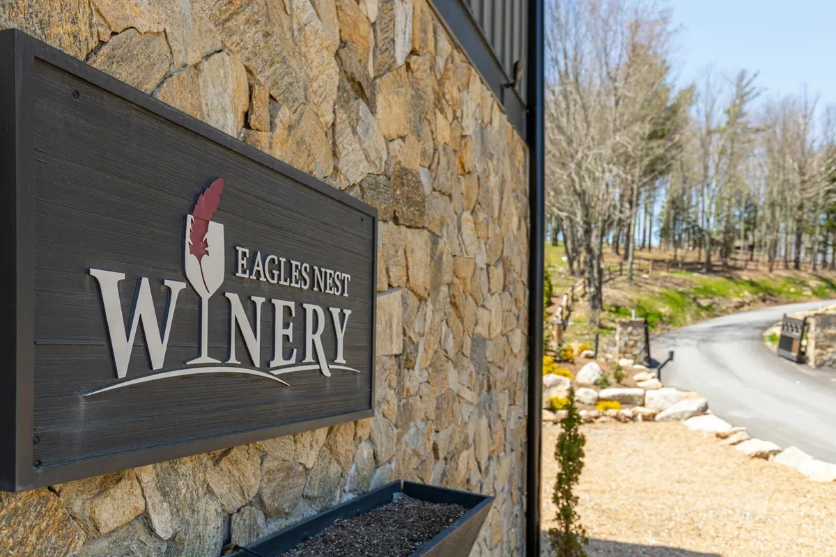 This exterior image features a welcoming winery sign set against a stone wall, with a pathway leading toward the winery building. The landscape hints at scenic views and a sense of anticipation for the wine tasting experience.