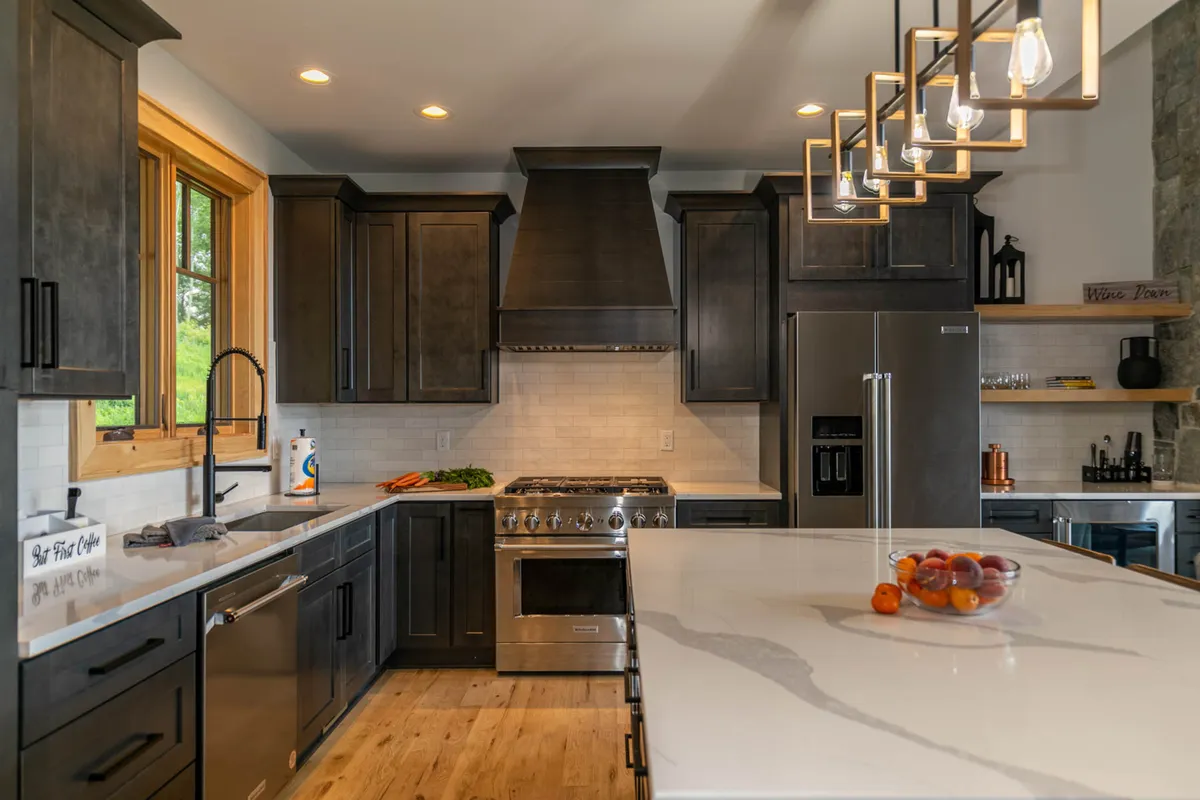 This image captures a spacious kitchen with a central island, stainless steel appliances, wooden cabinetry, and large windows bringing in natural light. A fruit bowl sits on the island, and the open layout hints at an inviting space perfect for cooking and entertaining.