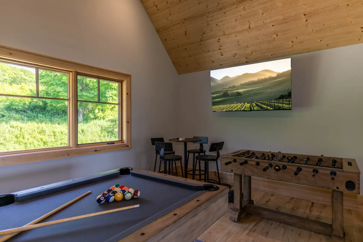 This inviting game room features both a pool table and a foosball table, a large window offering views, and a TV screen displaying a scenic landscape. The space is designed for group fun and relaxation, with plenty of room for movement and interaction.