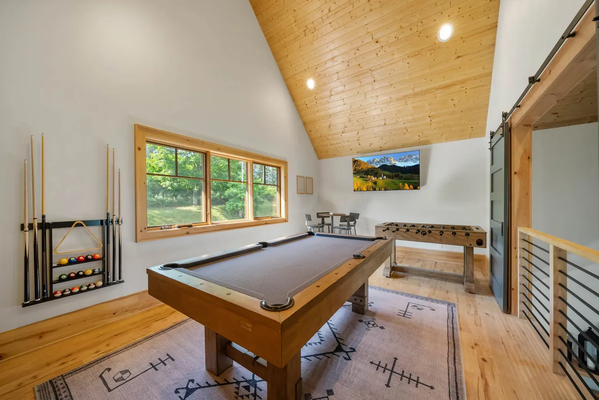 This image displays a spacious game loft featuring a pool table and a foosball table beneath a wooden ceiling. Large windows fill the room with natural light, and there is a TV with a landscape painting on the screen, creating a playful yet relaxed atmosphere ideal for friendly competitions and social gatherings.
