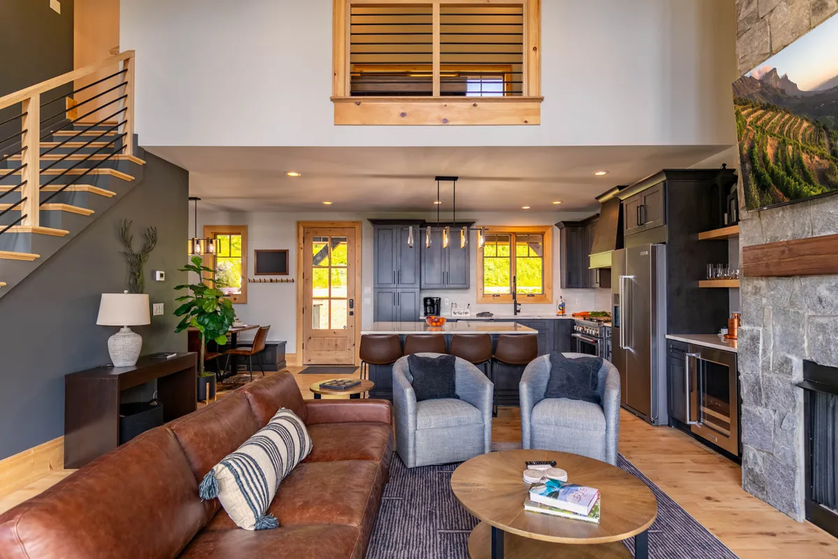 An airy, open-concept living space with a brown leather sofa facing a stone fireplace, a wooden dining table, large windows, and a staircase, seamlessly blending the great room and dining area for group activities and meals.