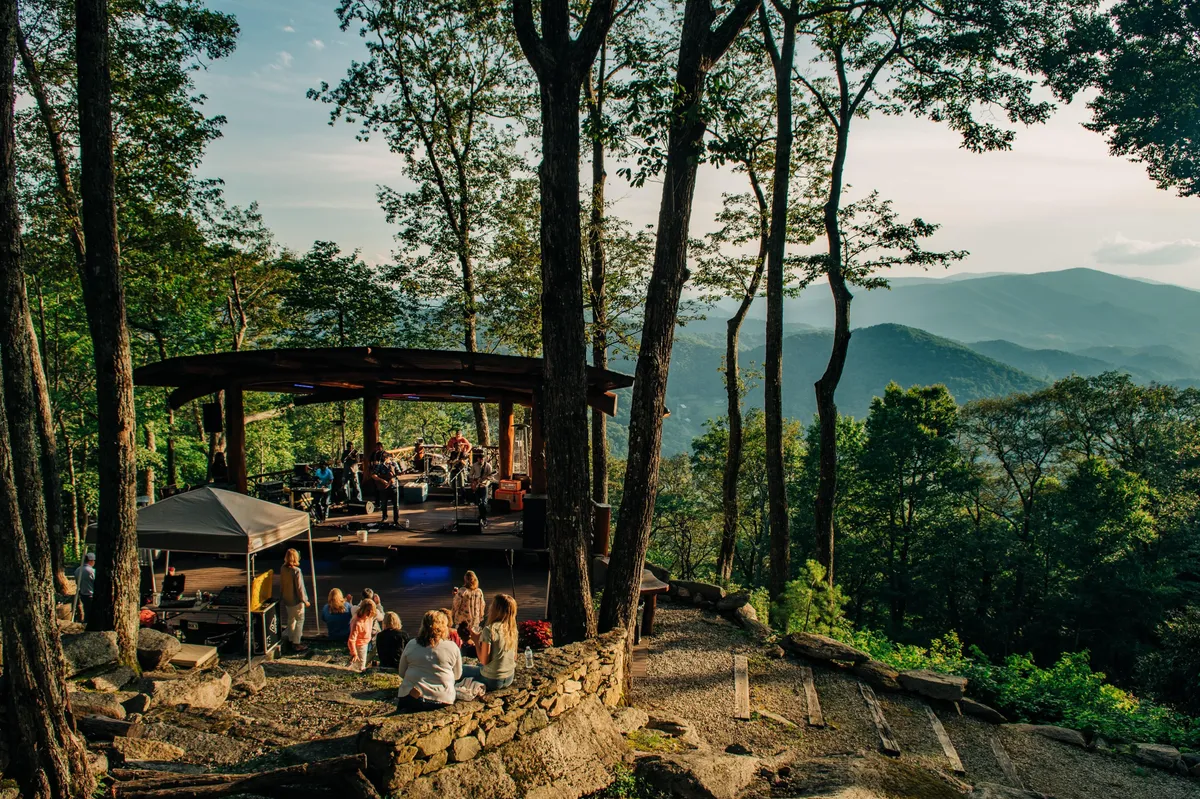 A vibrant outdoor amphitheater scene with a live music performance, audience members gathered together, and scenic mountain views beyond. The atmosphere is lively, communal, and full of opportunity for shared experiences under the sky.