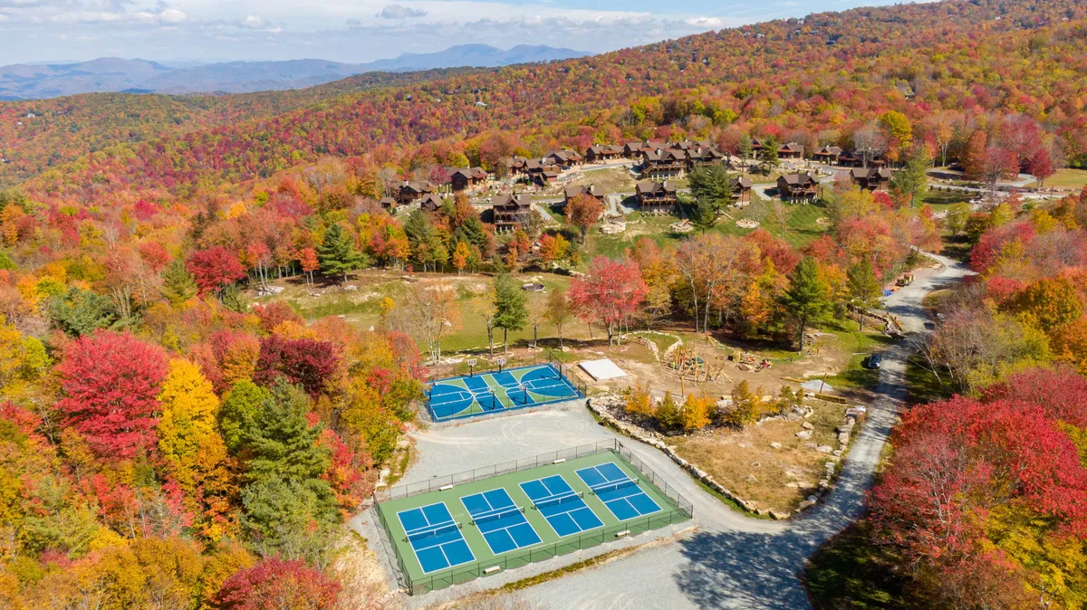 Colorful tennis courts nestled among vibrant autumn foliage, with residential buildings and forested hills in the background. The scene is energetic yet scenic, blending recreation with natural beauty.
