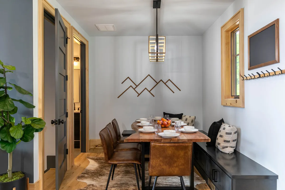 A stylish dining area featuring a long wooden table, a modern chandelier, mountain wall art, and a green plant, evoking warmth and togetherness for shared meals and celebrations.