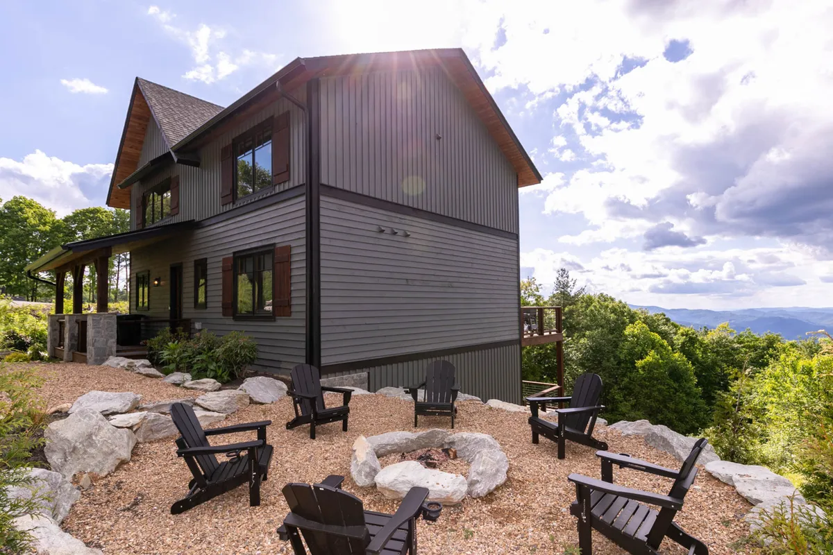 This image features a cozy exterior gathering space with a fire pit surrounded by Adirondack chairs, set against a backdrop of lush greenery and the house's facade. The inviting setup suggests a perfect spot for relaxing outdoors with pets and family after a day of mountain exploration.