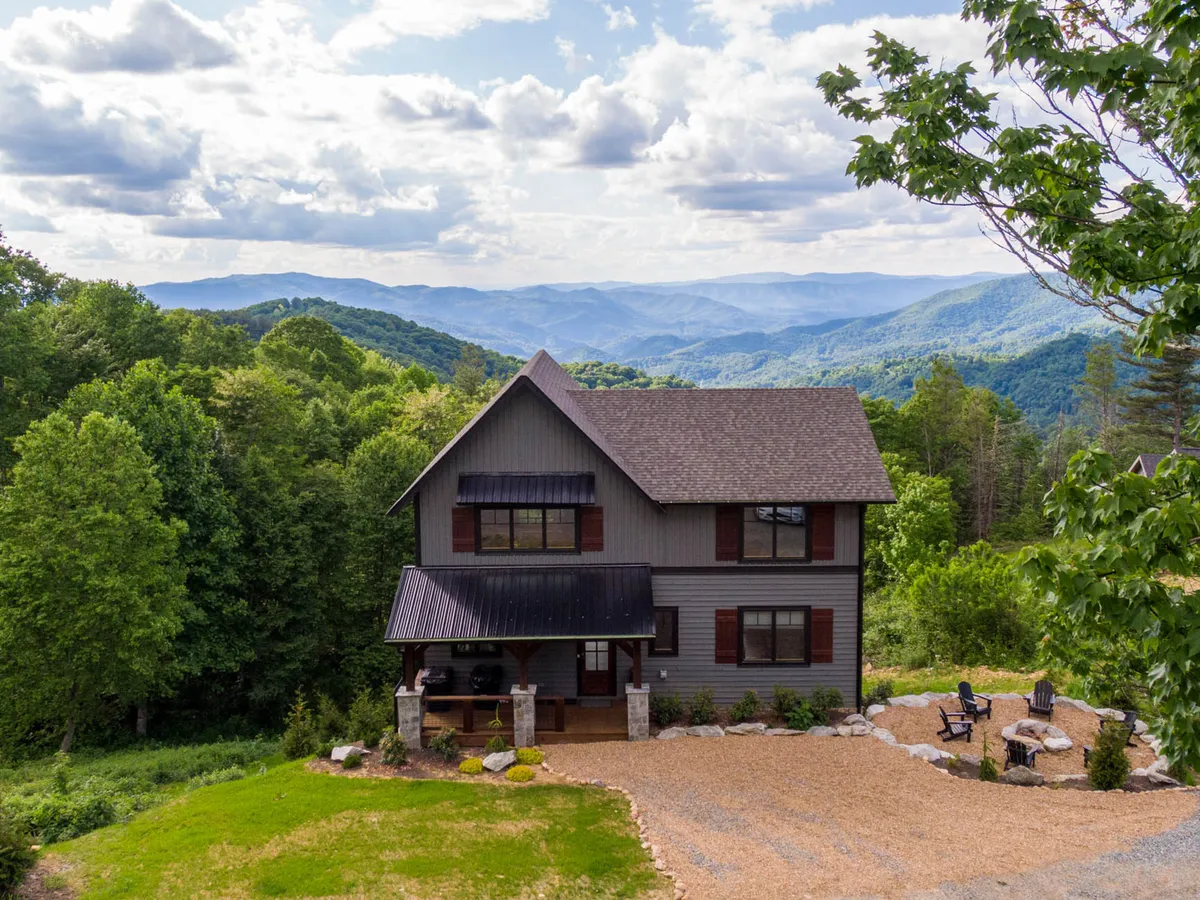 The image showcases a stunning, newly built single-family mountain home nestled on a landscaped hillside with inviting outdoor seating. The house overlooks sweeping mountain vistas, with lush gardens and a spacious porch—perfect for relaxing or entertaining while soaking up the tranquil scenery.